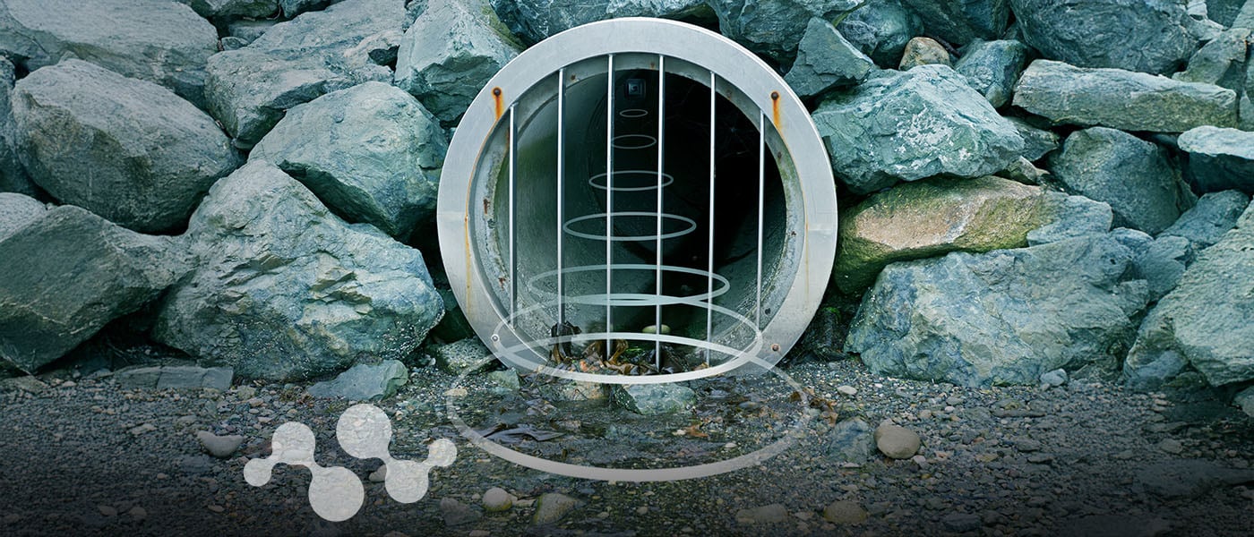 A circular drainage grate with vertical metal bars installed within a rocky shoreline or seawall. The drainage pipe extends into the rocks, showing water level markers inside. A small water molecule graphic appears at the bottom of the image. This represents an IoT-enabled smart drain monitoring system using LoRaWAN® technology for remote water level detection and flood prevention.